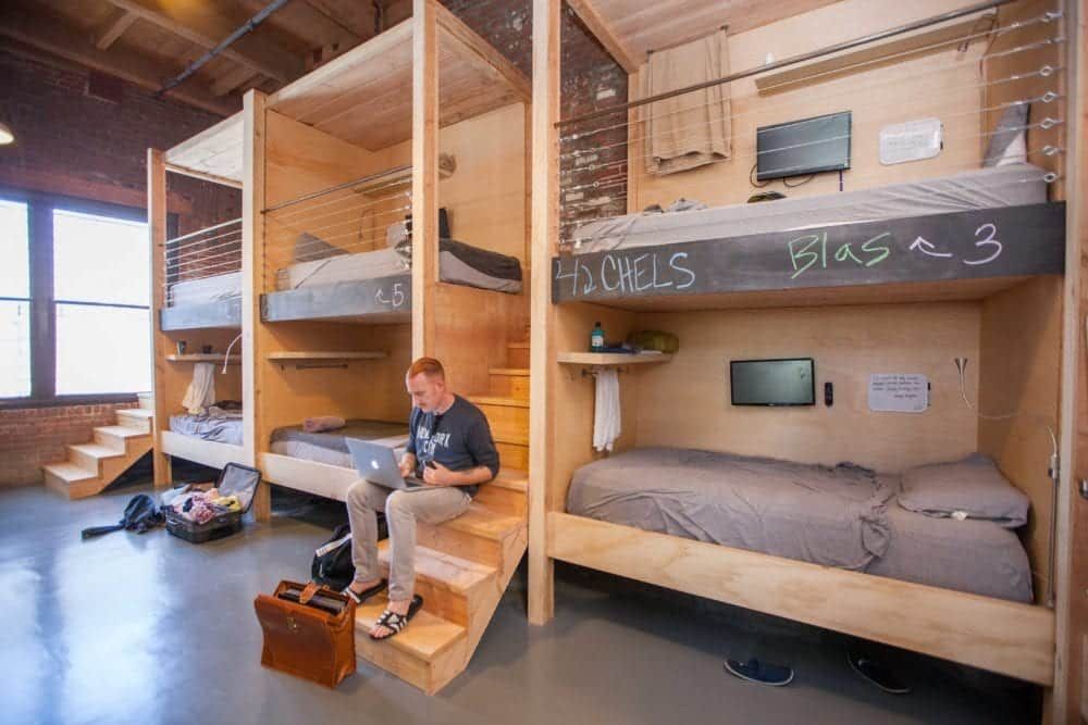 Man on steps using laptop in a pod-style bunk bed room. Wooden structures, exposed brick, bright light.