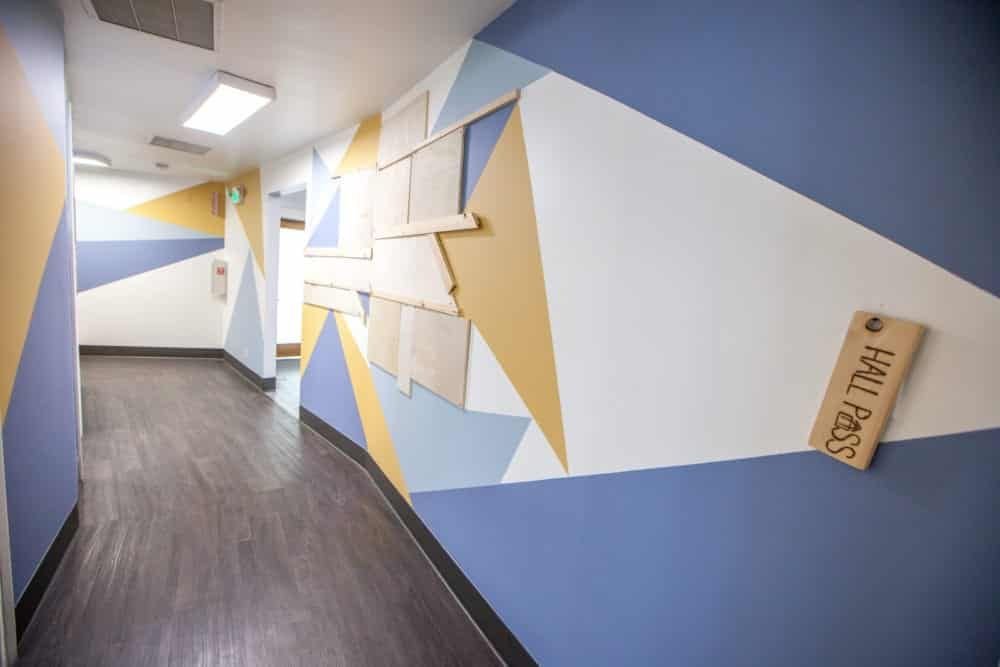 Hallway with geometric painted walls in blue, white, and yellow. Wooden hall pass hangs on the right.