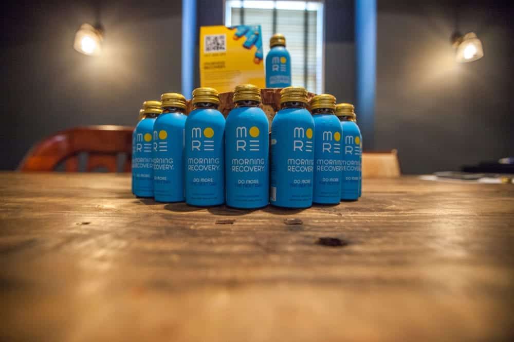 Blue bottles of "MORE" recovery drink on a wooden table, with a yellow box in the background.
