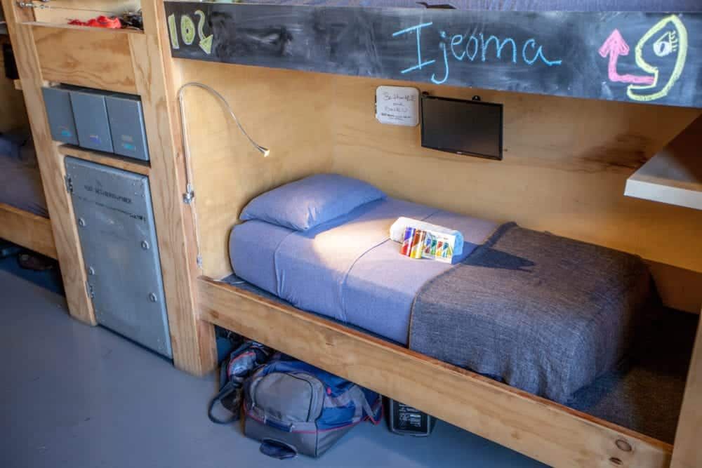 Bunk bed with a blue comforter, light, small TV, and storage. Name "Ijeoma" written on the wood.