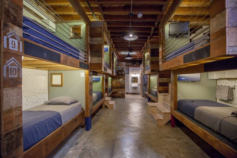 Bunk beds line a long hallway. Wooden frames with gray and blue bedding, built-in TVs.
