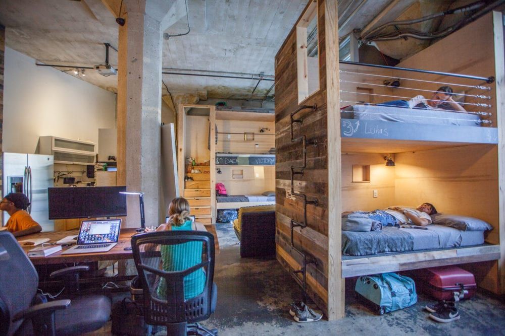 A shared living space with bunk beds, desks, and people working/resting in a warehouse setting.