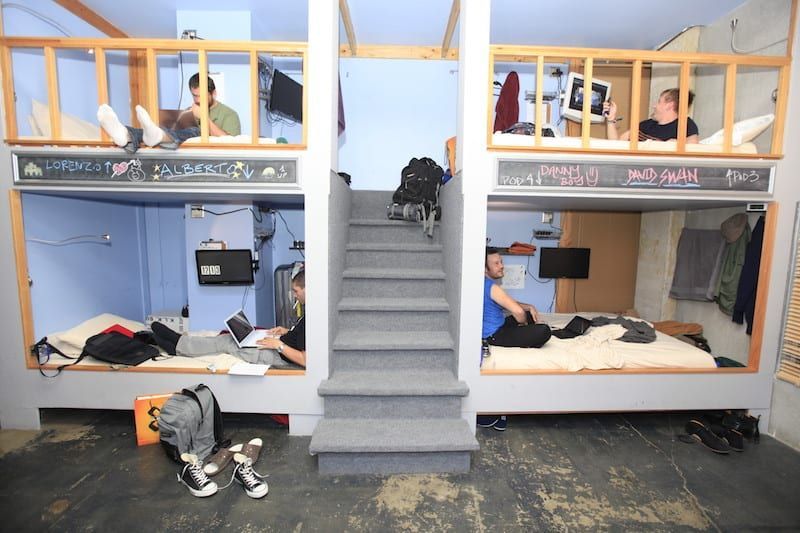Two-story bunk beds in a room; people on beds, some using computers. Grey stairs lead up.