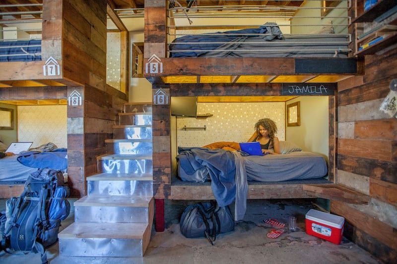 Cozy hostel room with wooden bunk beds, a woman using a laptop on the lower bunk, and a backpack nearby.