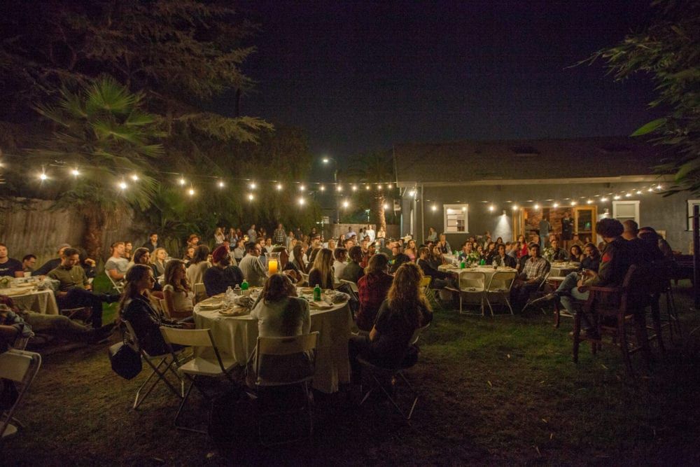 Outdoor night gathering with people seated at tables, lit by string lights, in a backyard.