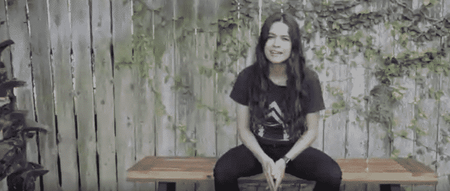 Woman sitting on a bench in front of a wooden fence, wearing black, speaking with foliage.