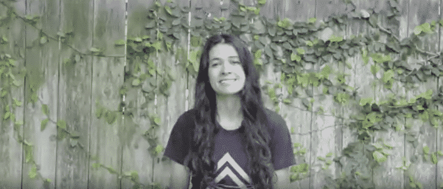 Woman with long dark hair smiles in front of a wooden fence covered in green vines.