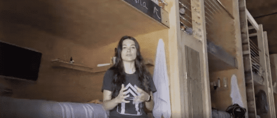 A woman with dark hair gestures in a room with bunk beds, likely a hostel.