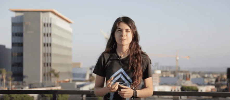 Woman with long hair standing outdoors with buildings in background, wearing a black t-shirt with a blue logo.