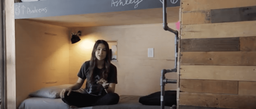 Young woman sitting in a bunk bed, looking at camera. A wall of wood planks and metal pipes is to the right.