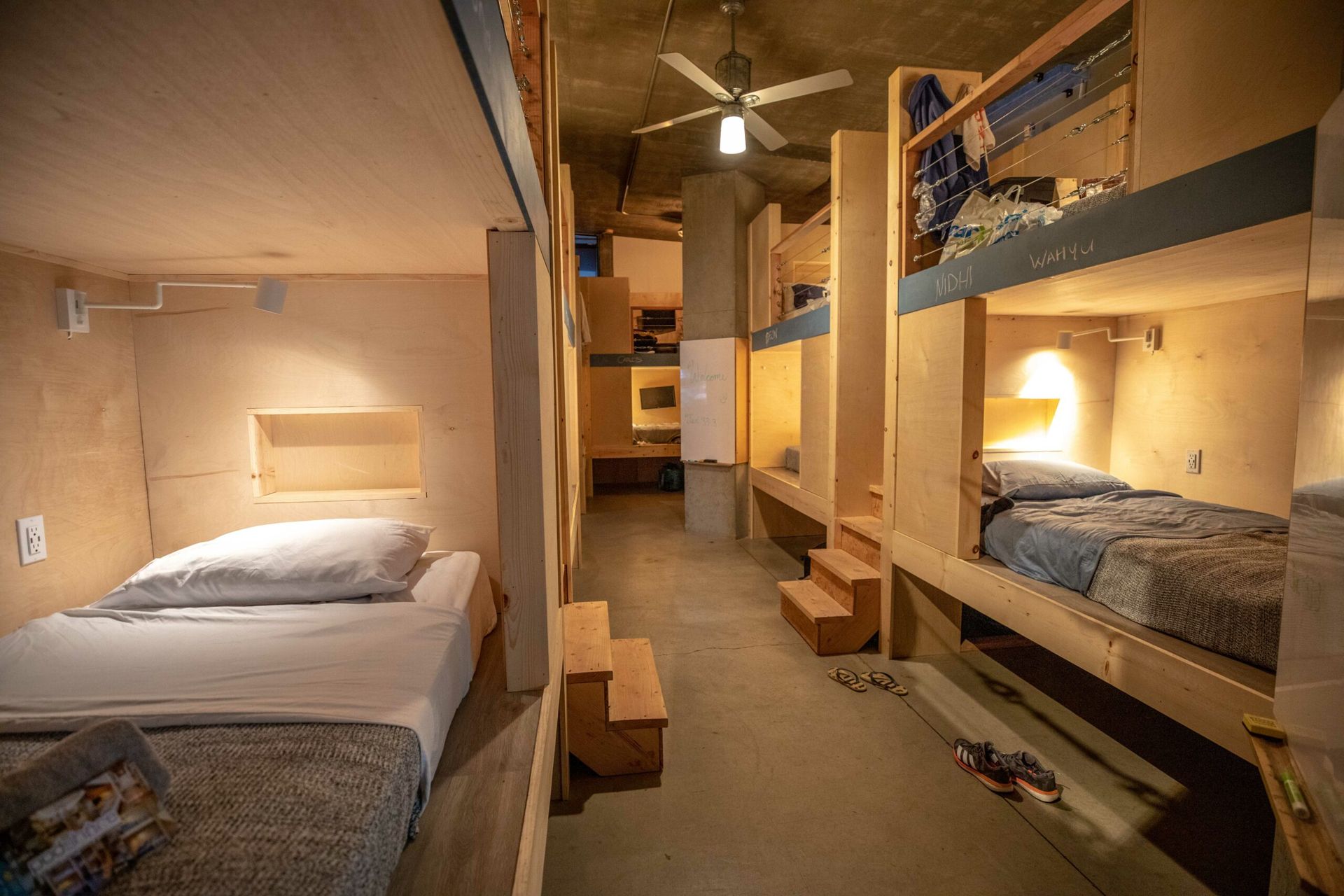 Hostel room with built-in bunk beds and single beds, concrete ceiling, warm lighting.