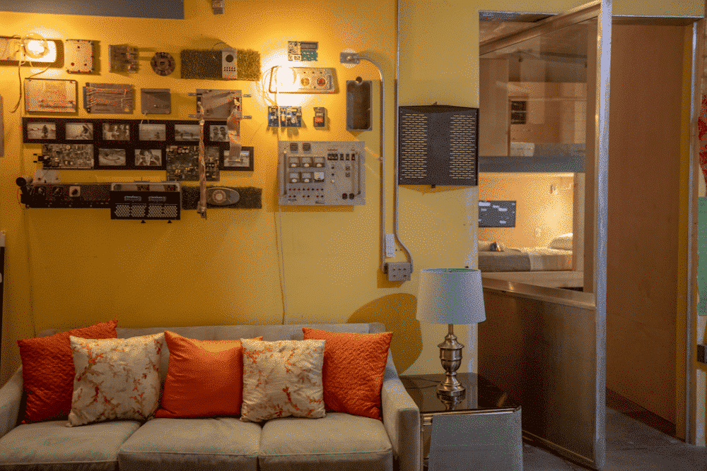 A cozy living room with a couch, orange pillows, and eclectic wall art; a door leads to a bright space.