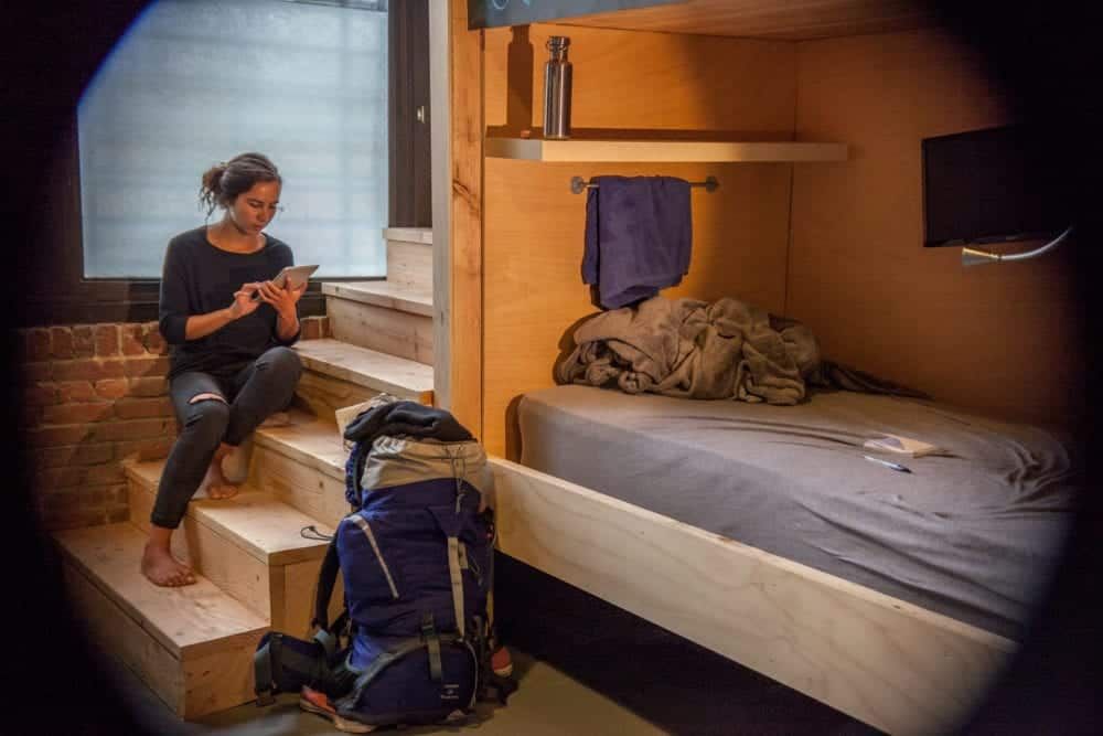 Woman sitting on stairs in a compact room, backpack nearby; bed and shelf visible.