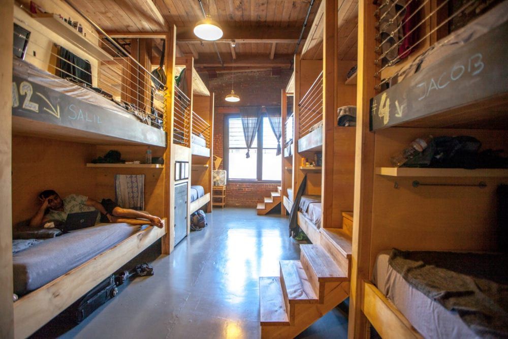 Hostel room with wooden bunk beds and a person working on a laptop in one of the beds.