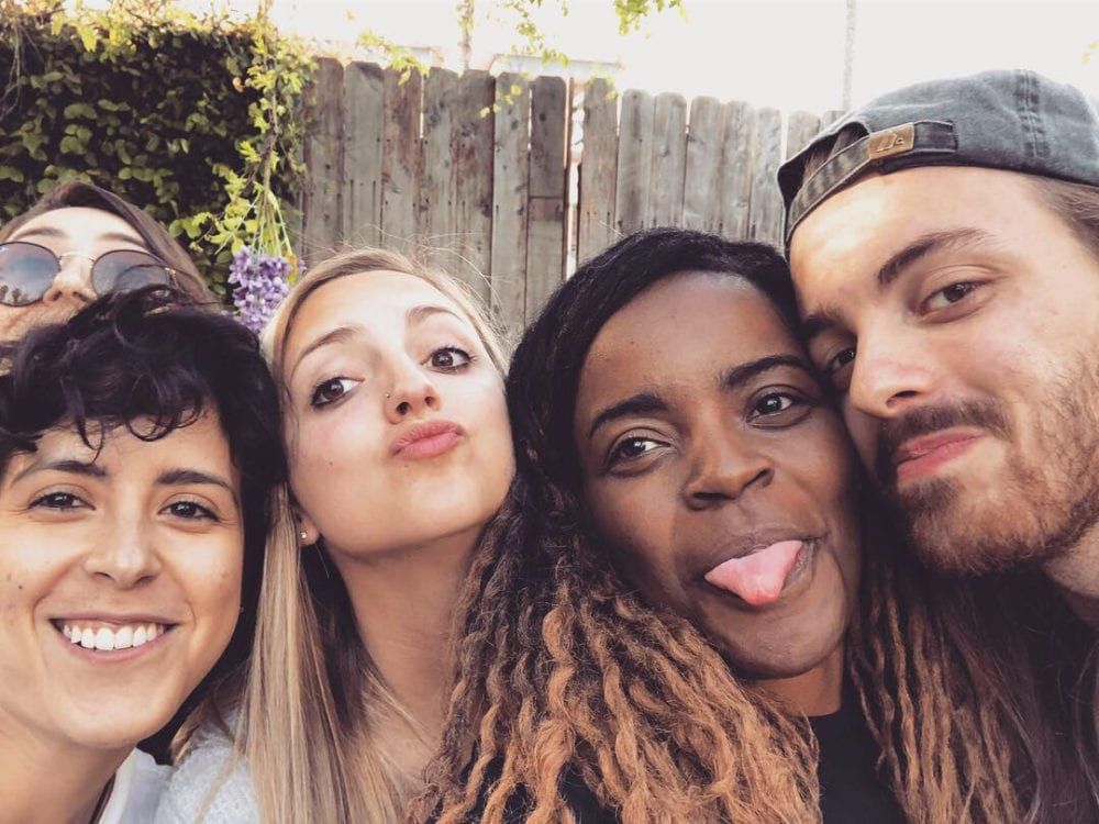 Group selfie with five diverse friends making silly faces outdoors by a wooden fence.