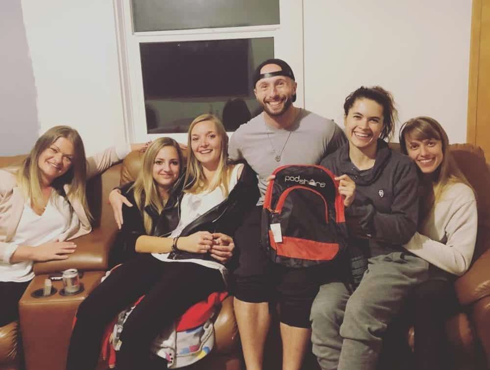 Group of six people smiling, posing on a brown couch, holding a backpack in a cozy indoor setting.