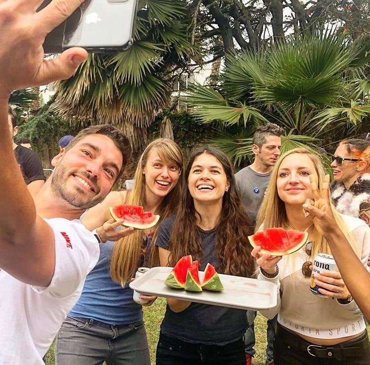 Group of friends taking a selfie with watermelon outside. Smiling, sunny day, casual attire, and food.