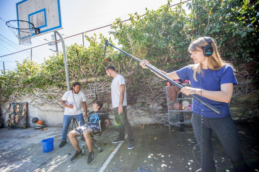 A film crew records on a basketball court. A boom operator holds a microphone while actors stand by.