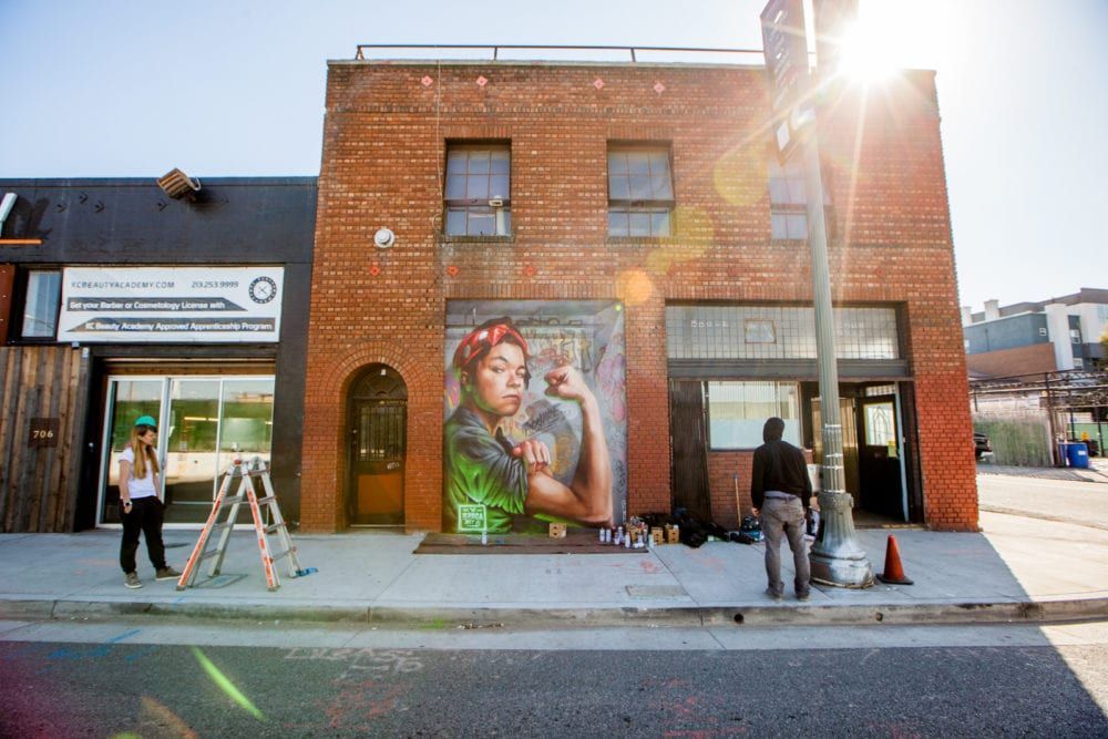 Mural of a woman flexing arm on a brick building. Artists standing on sidewalk. Sunny day.