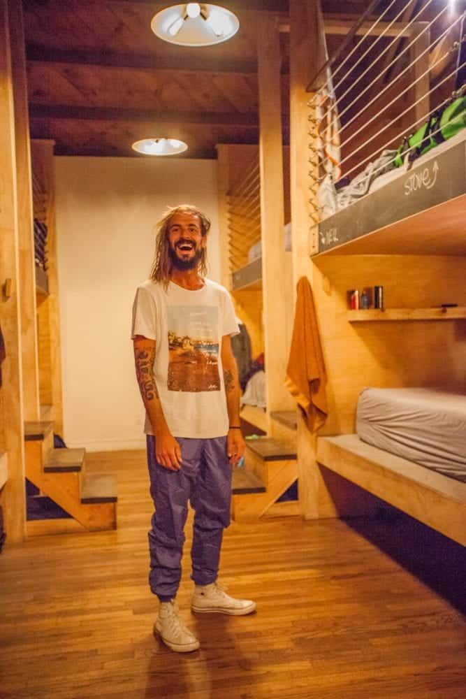 Man in a hostel smiling, standing between wooden bunk beds. He wears white shirt and purple pants.
