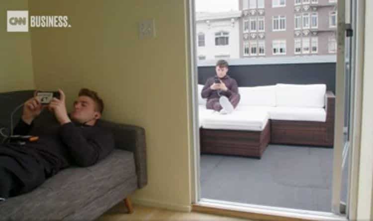 Man on couch with phone, another on outdoor sofa. Modern apartment, open doorway, buildings in the background.