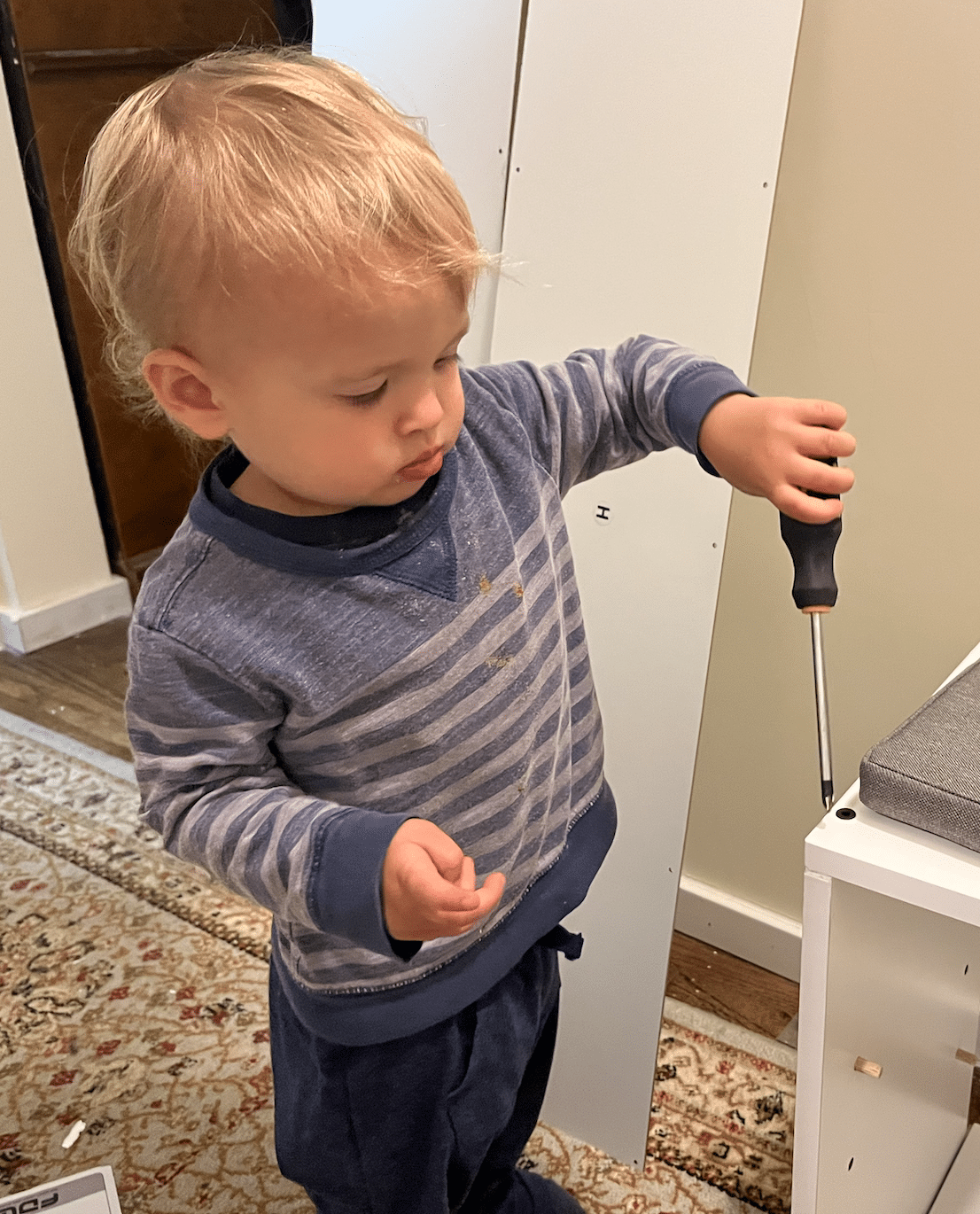 Blonde toddler in blue striped shirt using a screwdriver on furniture. Inside.