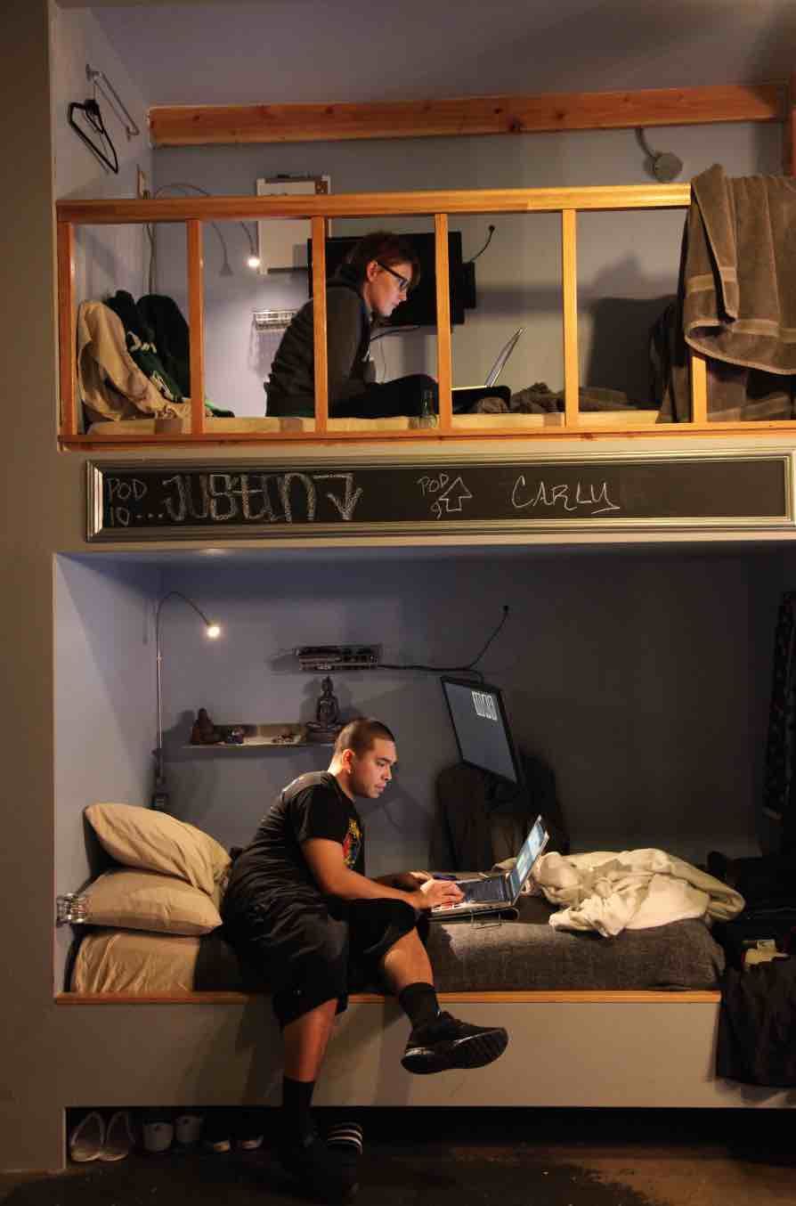 Two people in a cozy loft-style room. One works on a laptop on a lower bed, the other on a higher platform.