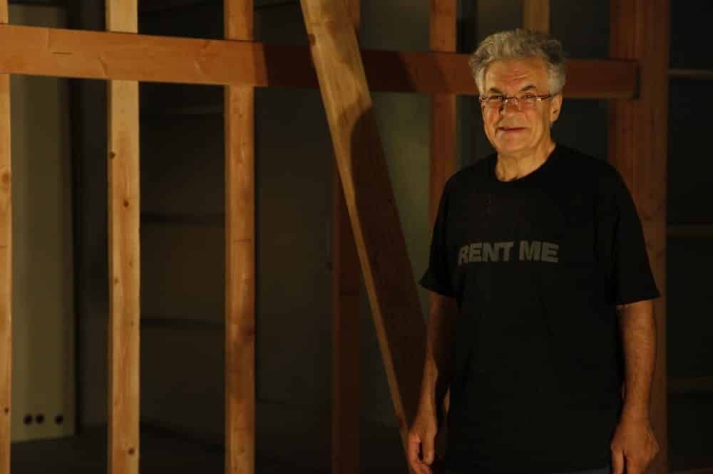 A man with gray hair and glasses stands in front of a wooden wall frame; he wears a black shirt that says 