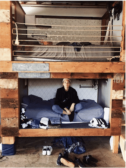 Person sitting in a bed within a wooden bunk bed structure. Clothes and shoes scattered below.