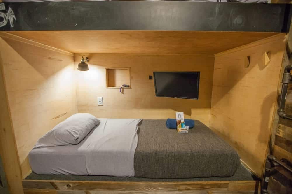 Wooden bed in a small, cozy cubicle with TV, lamp, and bedding.