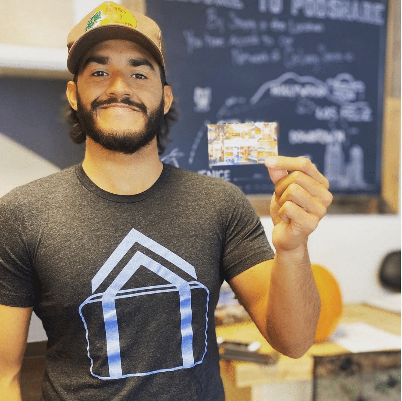 Man holding business card; wearing hat and grey shirt. Smiles in front of a chalkboard.