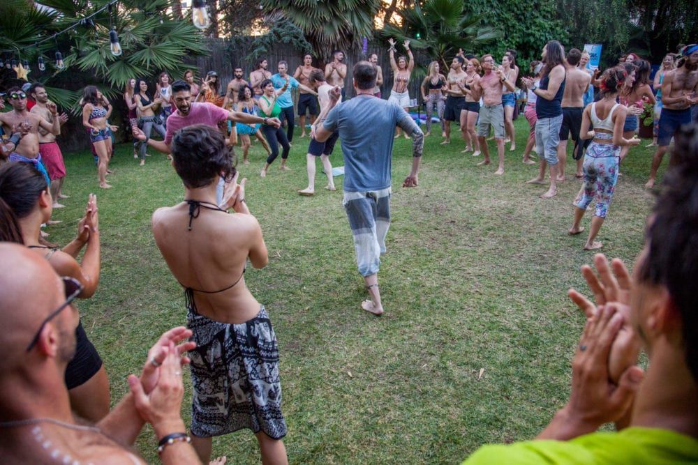 People dance in a grassy outdoor area, some clapping, in a lively, informal gathering.