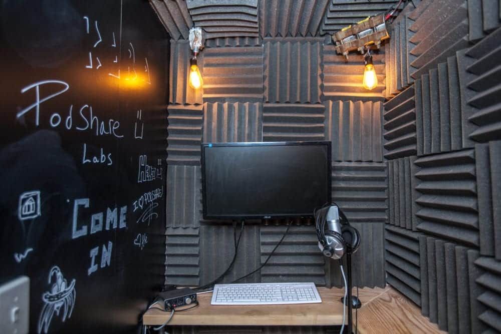 A soundproof podcasting booth with a desk, microphone, screen, and acoustic panels.