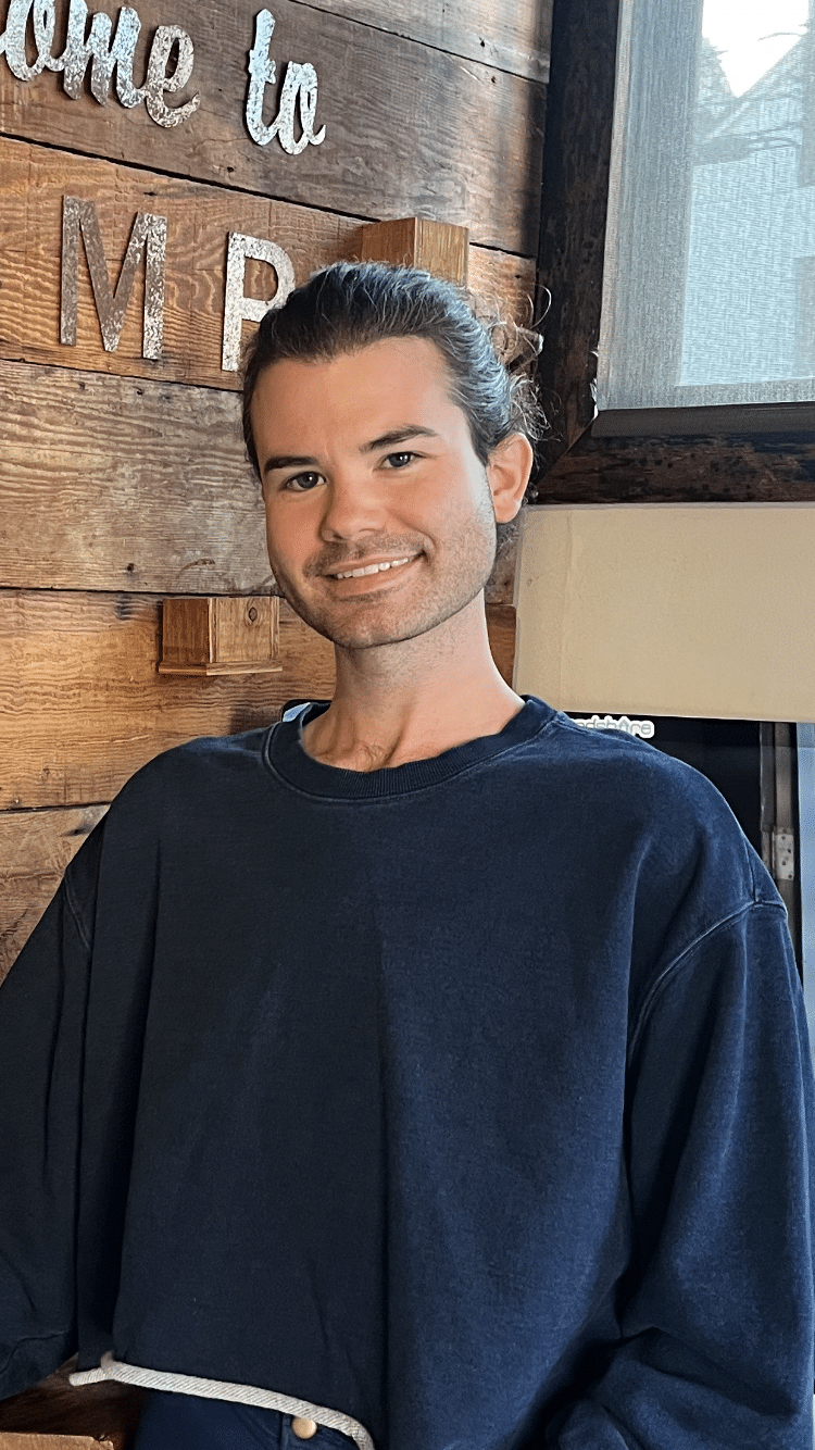 Man with dark hair smiling in front of a wooden wall, wearing a blue sweatshirt.