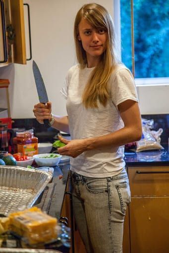 Woman in kitchen holds knife, looks at camera. Wearing white shirt, jeans. Cooking ingredients on counter.