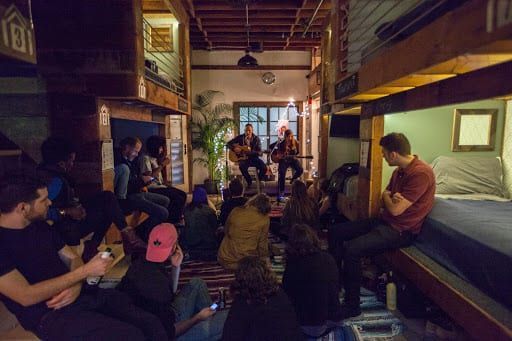 People listening to musicians playing guitars in a room with bunk beds. Dim lighting.
