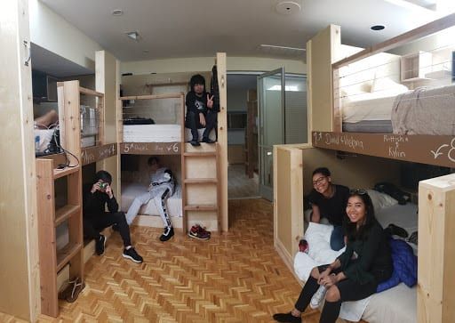 A group of teens in bunk beds in a room, some smiling. Wooden walls, parquet flooring.