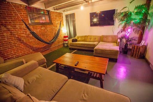 Cozy lounge with sectional couches, a hammock on a brick wall, and a coffee table, bathed in colorful ambient light.