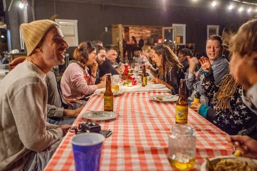 People at a long table with red checkered tablecloth, eating and drinking outdoors at night.