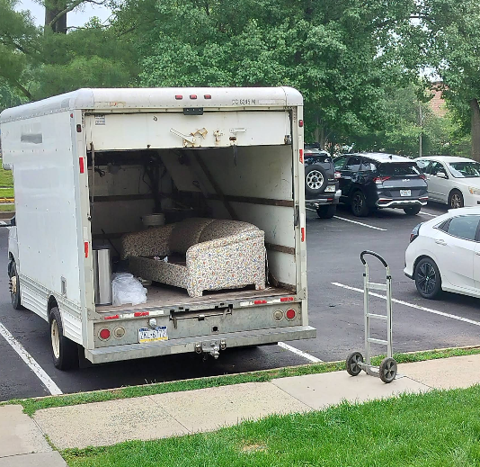 Moving truck with open back in parking lot. A couch and other items are inside. Hand truck visible.