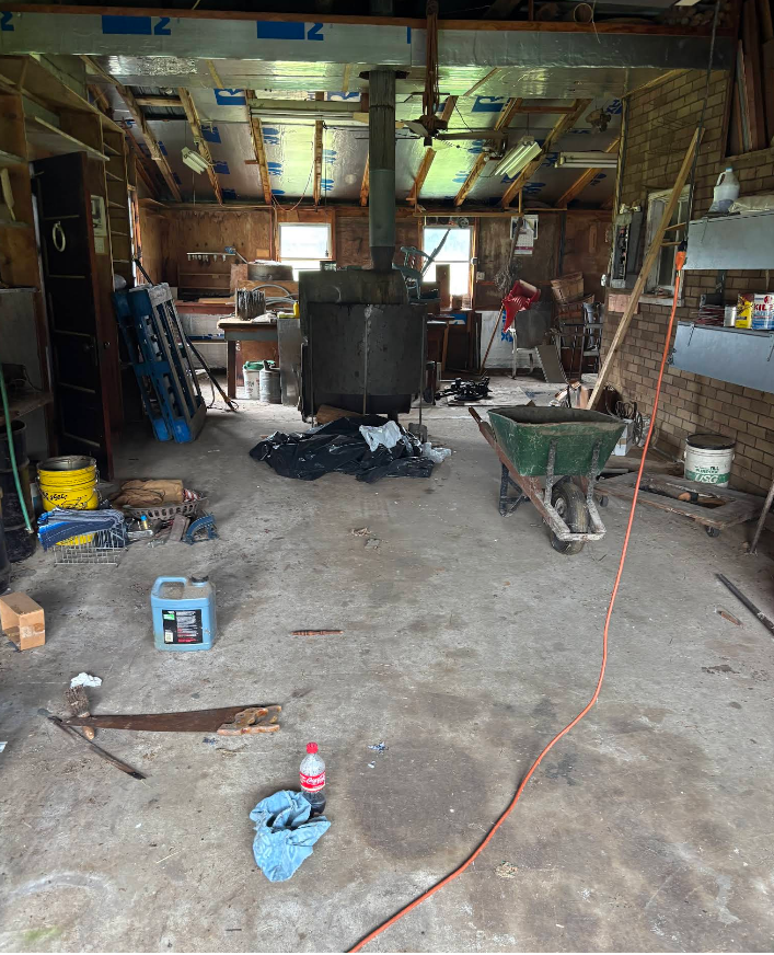 Messy workshop interior with a large central industrial machine, tools, and materials scattered on a concrete floor.