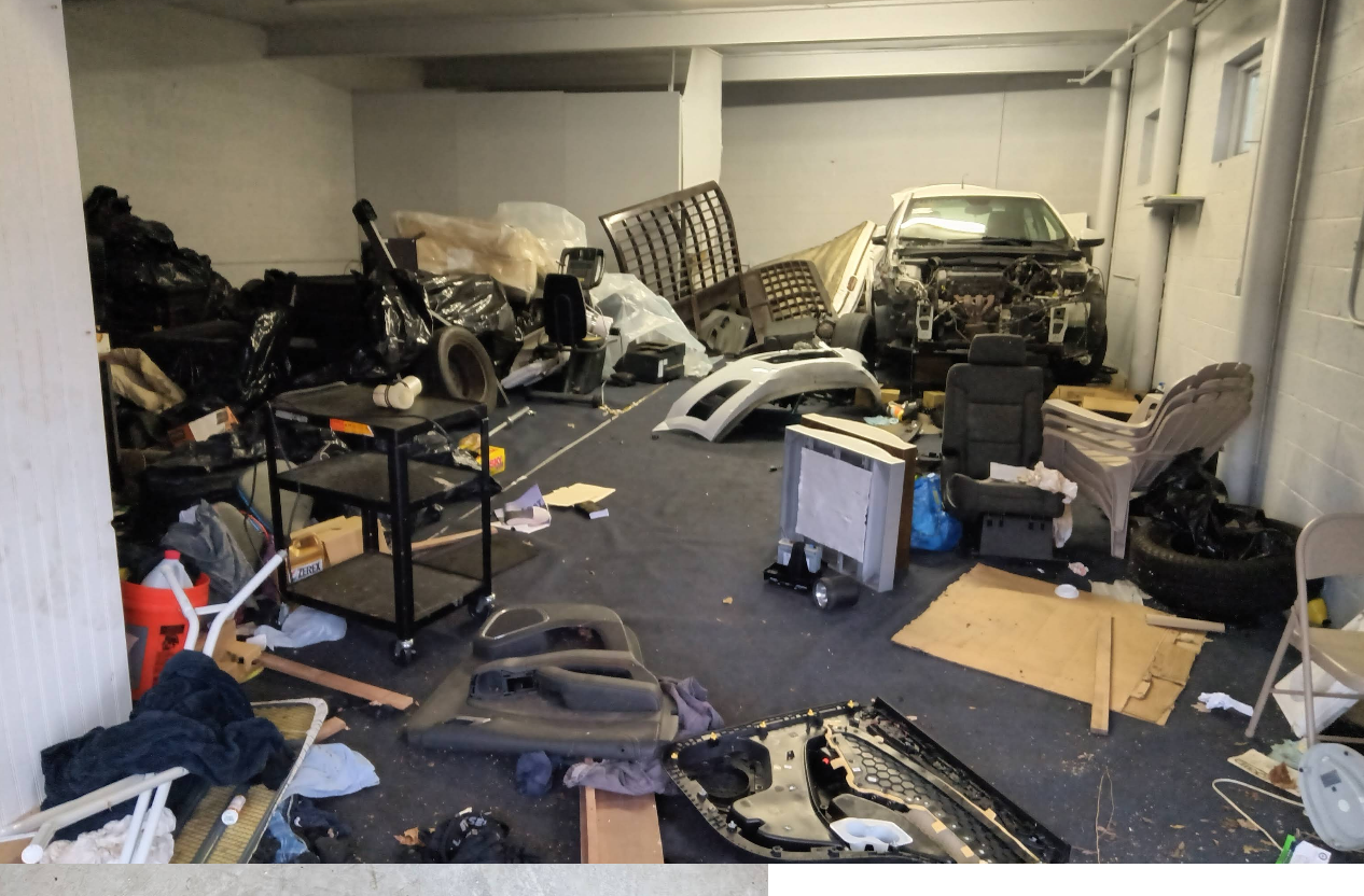 A cluttered garage with car parts and debris, including a car missing its front.