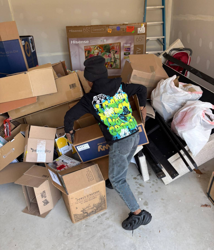 Person surrounded by moving boxes in a cluttered garage, wearing a black hoodie with a colorful design.
