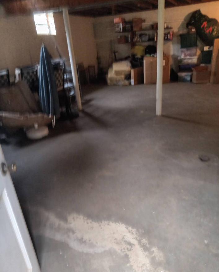 Basement with concrete floor and clutter.  White door in foreground. Window in background.