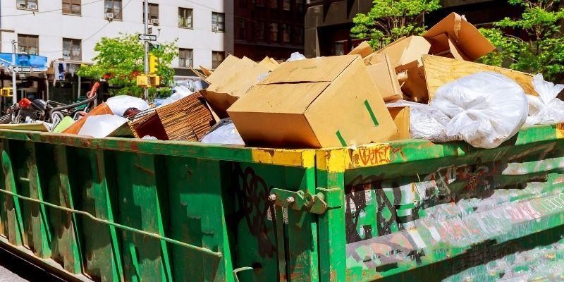 Green dumpster overflowing with cardboard boxes and trash on a city street.