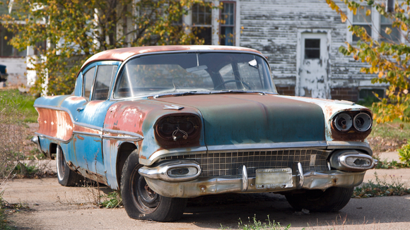 Rusty, vintage blue and green car, parked in front of a dilapidated white house.