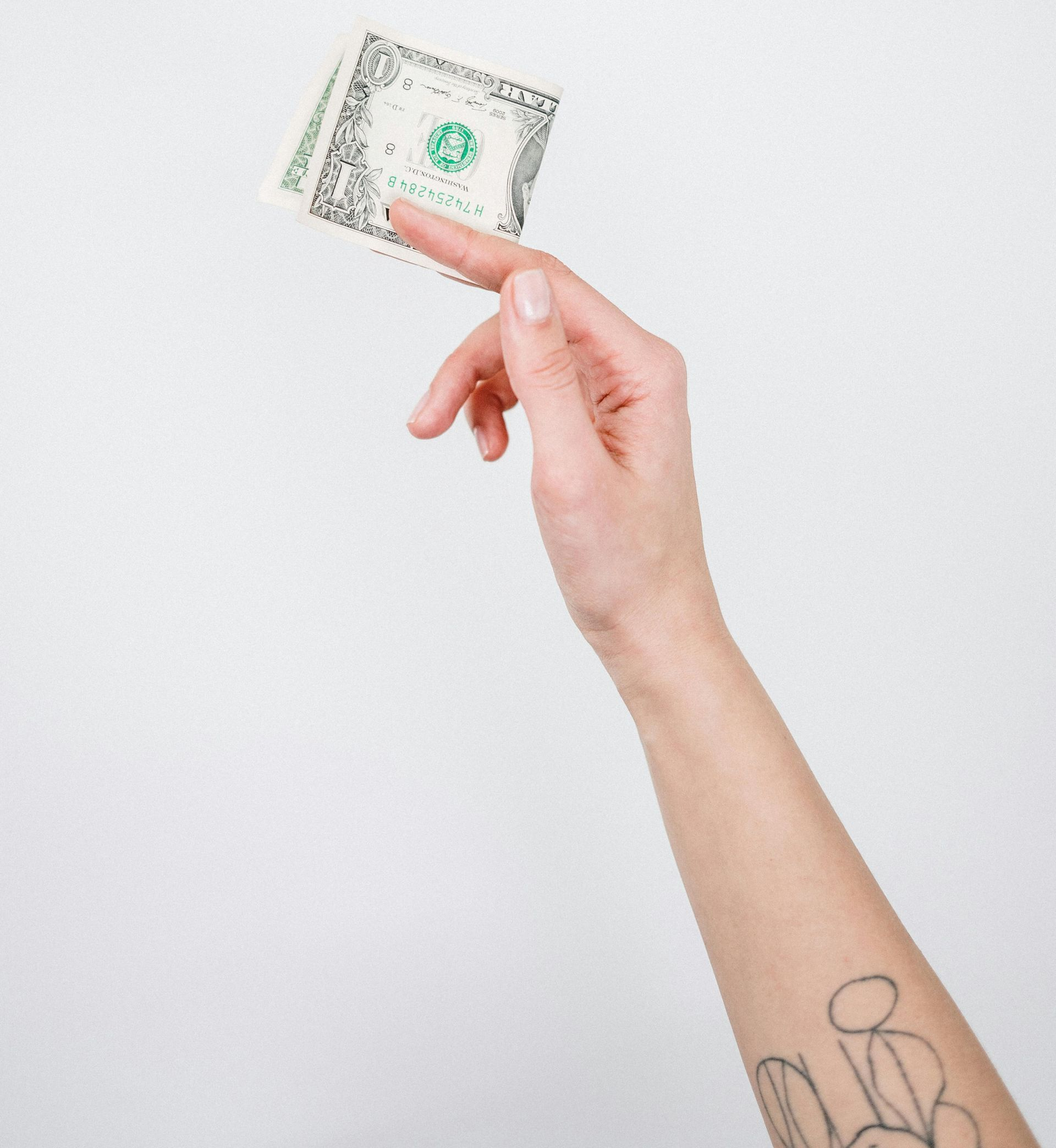 Hand holding two one-dollar bills against a white background. Arm has a small tattoo.