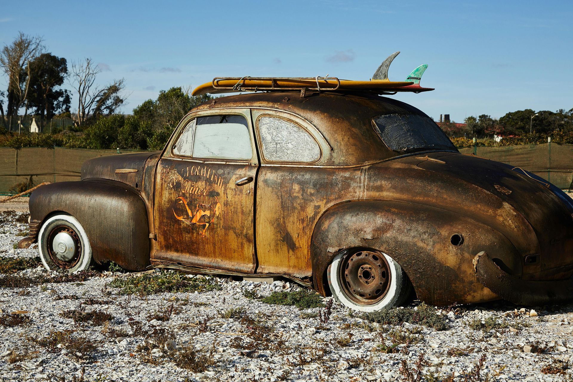 Rusty vintage car with surfboard on top, parked outdoors on gravel.