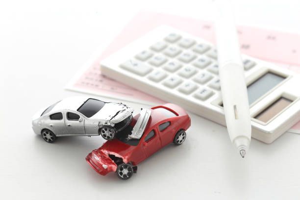 A silver toy car rests on top of a crushed red toy car next to a calculator and a pen on a white surface.