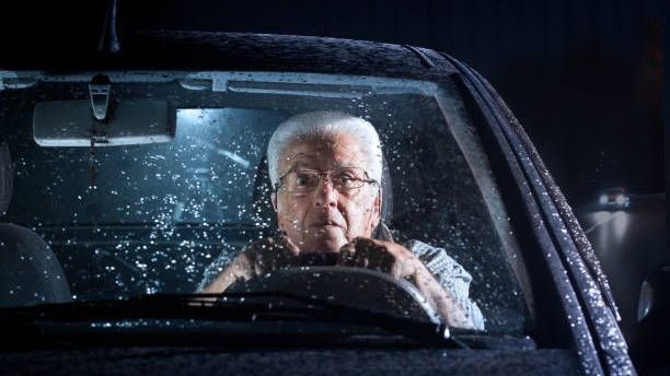 An older person with white hair and glasses driving a car at night in the rain, viewed through the windshield.